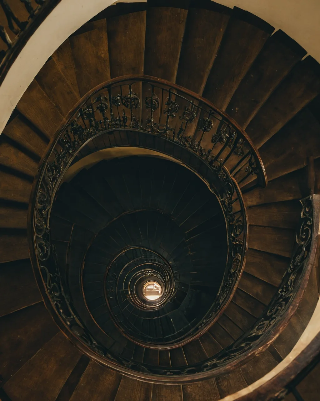 A dark wooden spiral staircase with ornate iron railings spiraling downward toward a glowing light.