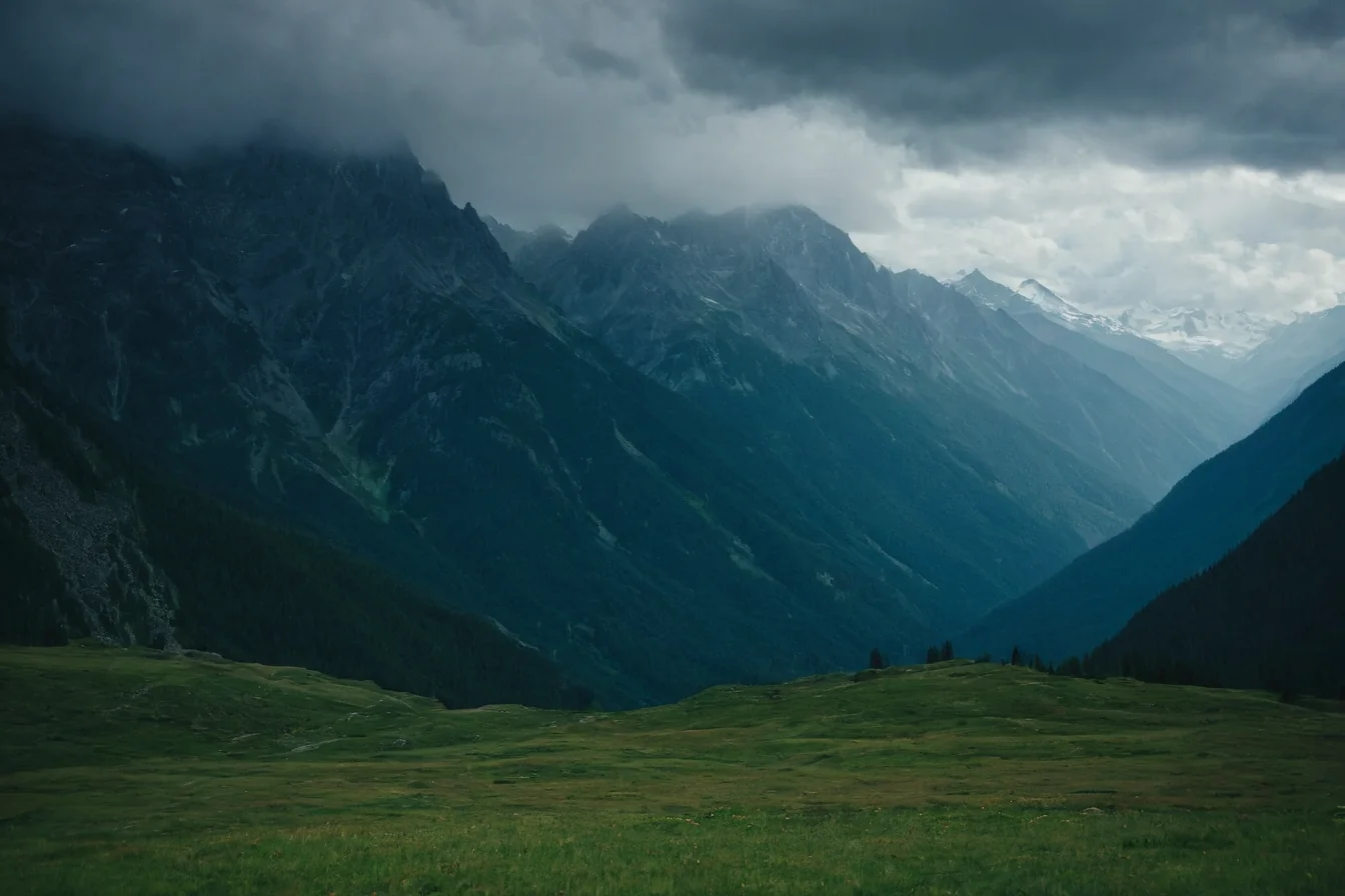 A wide green meadow with steep forested mountains rising into a cloudy sky.