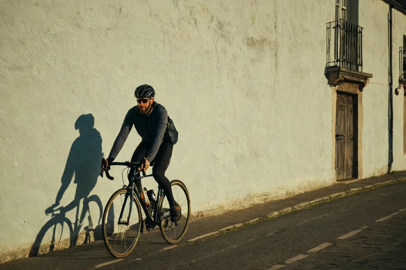 Cyclist riding a road bike on a cobblestone street with a strong shadow on a white wall.