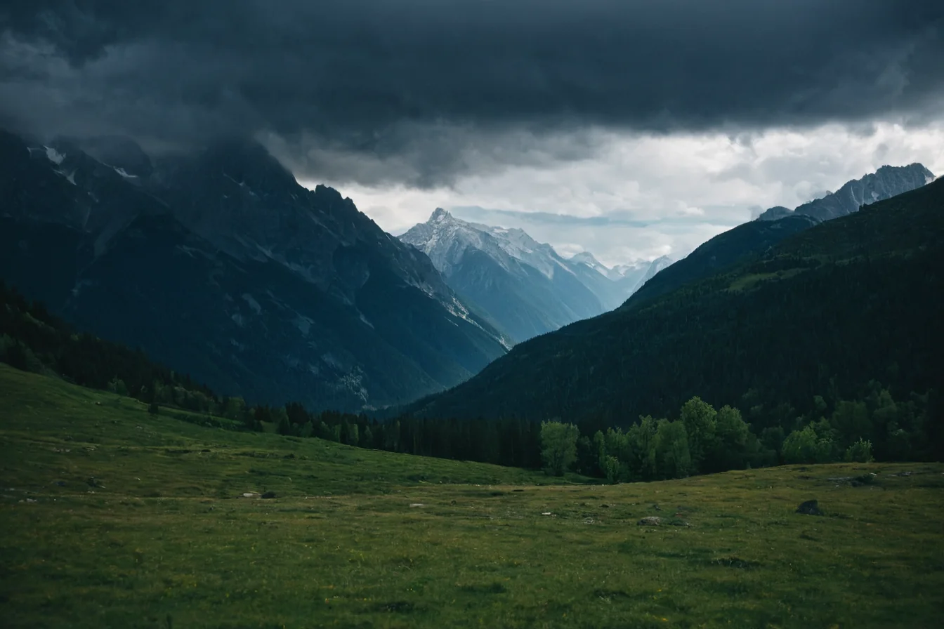 Dark storm clouds hover over snow-capped mountains and a green valley with trees.